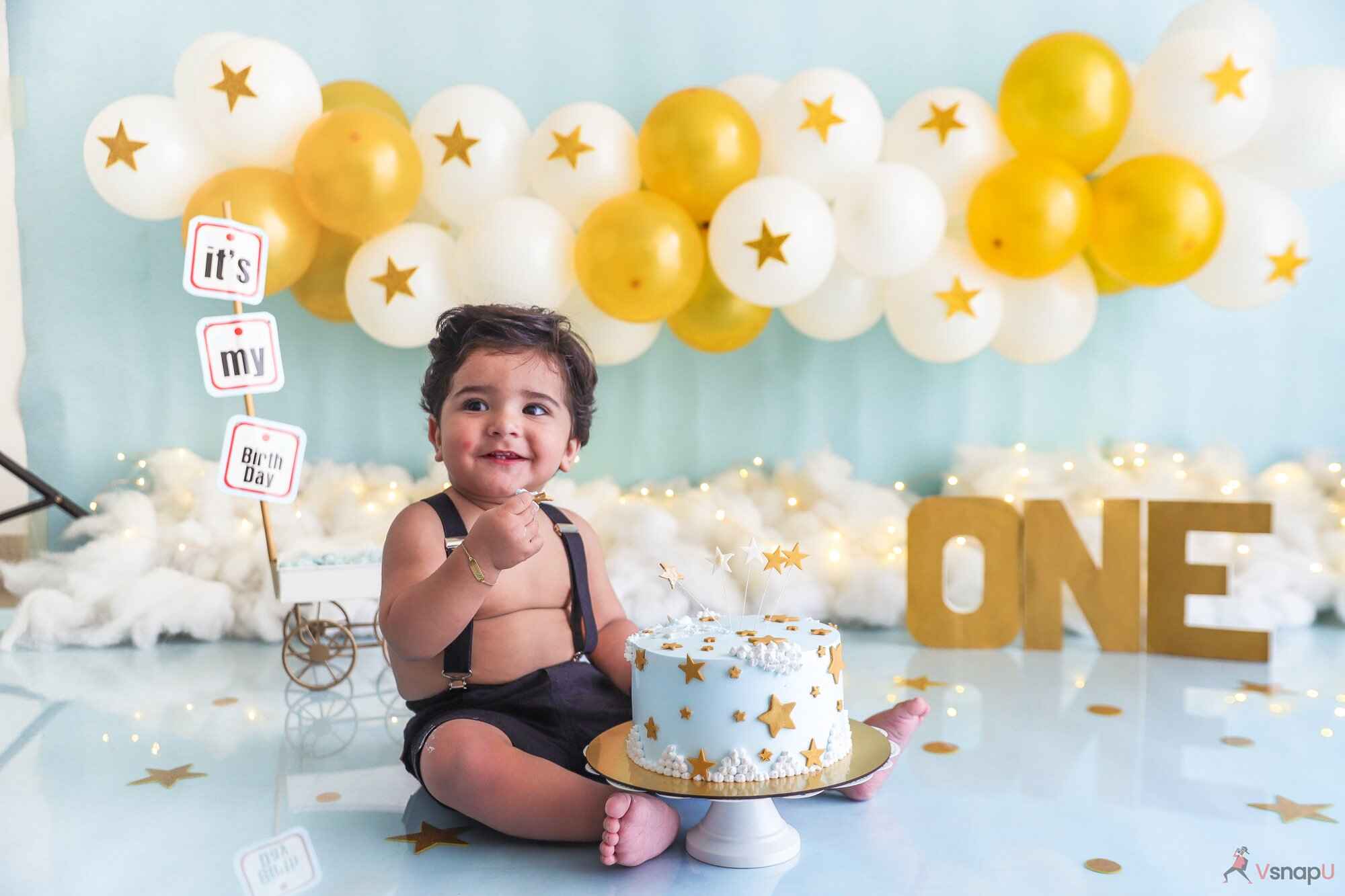 A one-year-old baby with a radiant smile, sitting with golden and white balloons during their baby photoshoot.